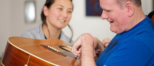 A support worker smiles as a man with disability plays guitar and smiles. They sit in a room.