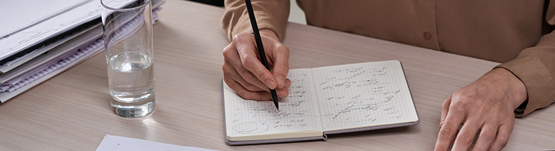 A hand writes with a pen in a notebook. Other papers are spread around the table
