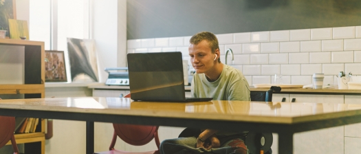 A young man with disability sits at a kitchen benchtop and looks at a laptop while wearing headphones.