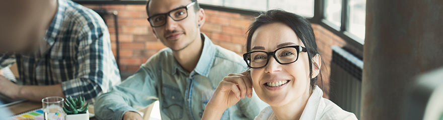 Two people in an office look up and smile. They both wear glasses. There is a brickwall and windows behind them.