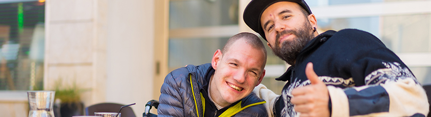 Two men sit outside. One man is using a wheelchair and smiling. The other man smiles and gives the camera a thumbs up.