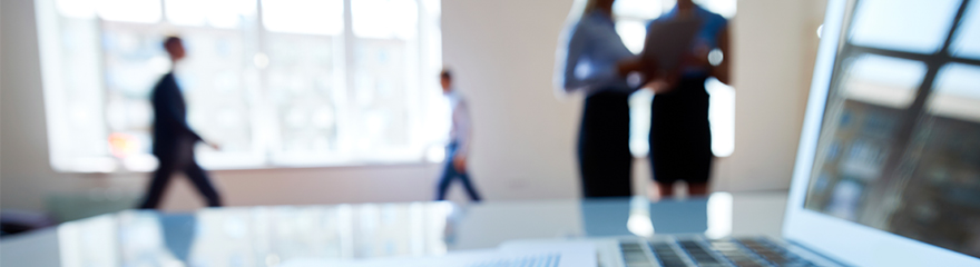 An out of focus image with a laptop in the foreground and office workers walking in the background