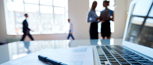 An out of focus image with a laptop in the foreground and office workers walking in the background