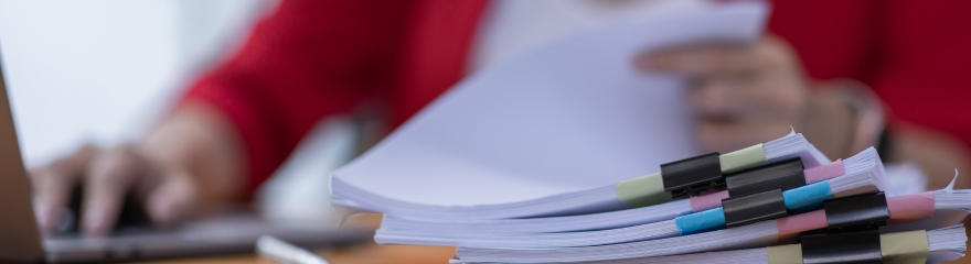 Close up of a stack of document with multicoloured bull clips, a laptop sits in the background