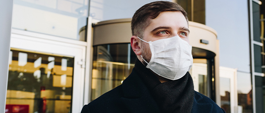 A person stand in front of a city building wearing a surgical mask