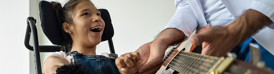 A young person in a powered wheelchair laughs along with their carer who is playing the guitar.