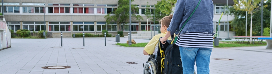 Support person guides a wheelchair away from the camera in an outdoor setting surrounded by building